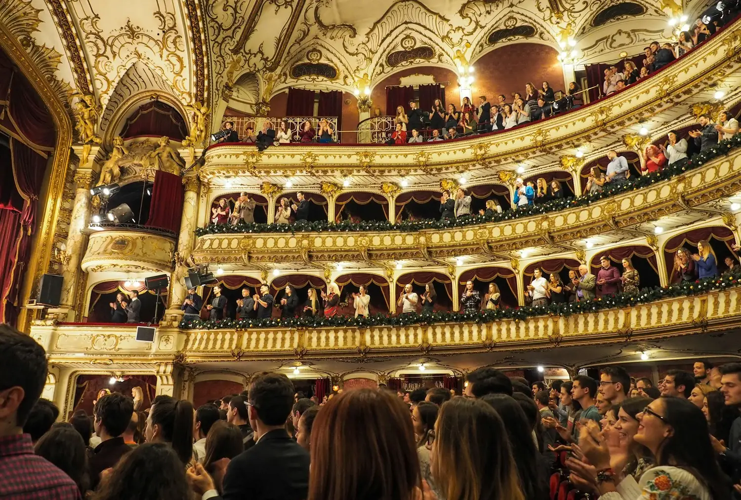 The ornate, gilded auditorium of a classical opera house, filled with an audience.